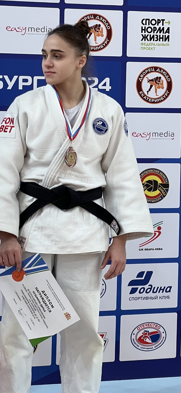 Frau in weißem Judoanzug mit schwarzem Gürtel auf einem Podest, die eine Medaille und ein Zertifikat hält, mit einem Logo-Plakat im Hintergrund.
