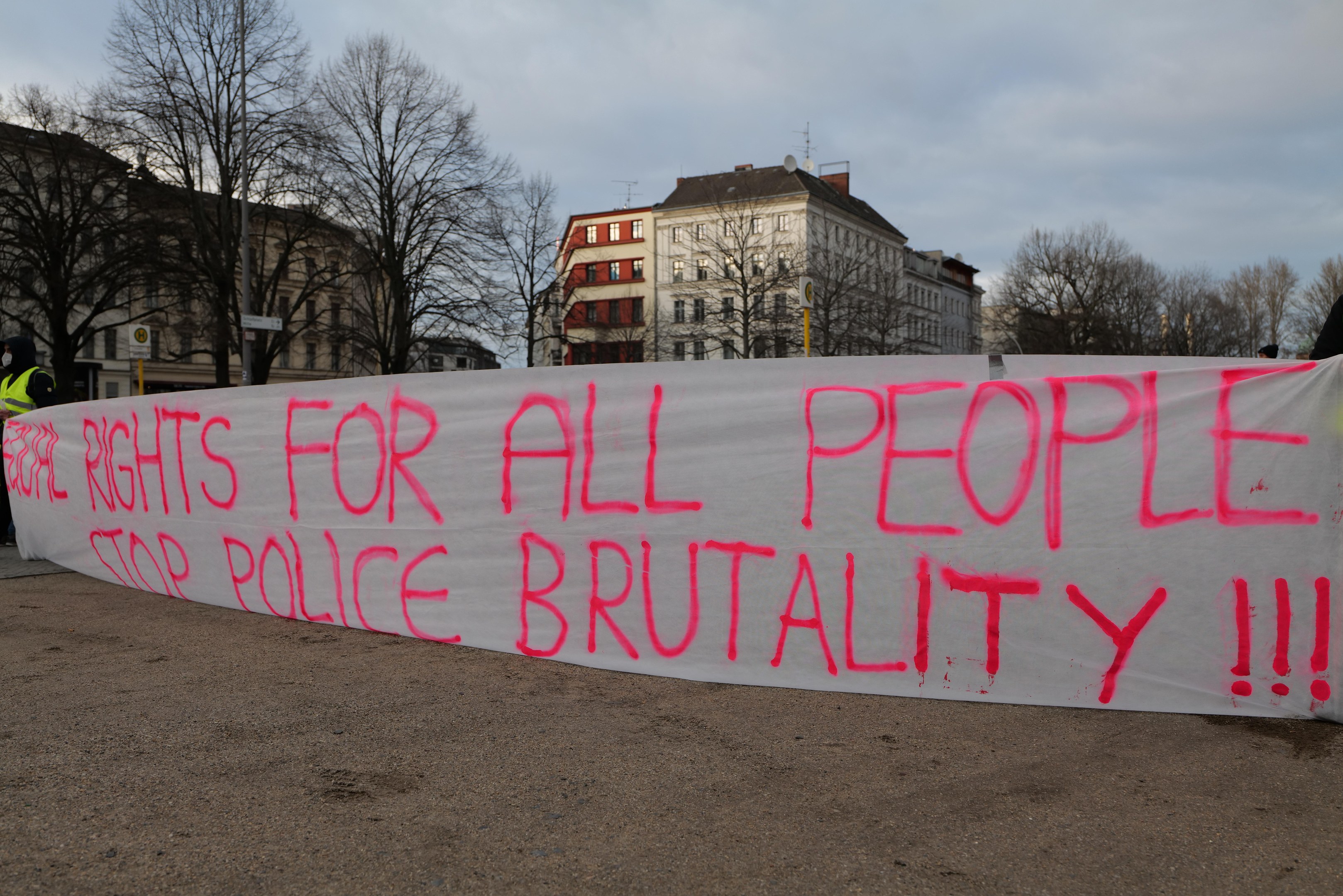 Group of people holding a banner reading "Rights for All People Stop Police Brutality" in front of a street pole, signboard, trees, buildings, and a cloudy sky.