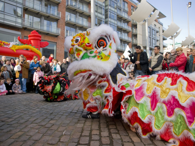 Lebendiges chinesisches Neujahrsfest in Amsterdam mit einem Löwen tanzen im Vordergrund und einer Menschenmenge drumherum, einige halten Kameras, vor einem Hintergrund aus Gebäuden, Laternenmasten und einem klaren blauen Himmel.