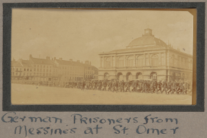 Eine Gruppe deutscher Kriegsgefangener aus Messines vor einem Gebäude unter einem klaren blauen Himmel, mit Text am unteren Bildrand.