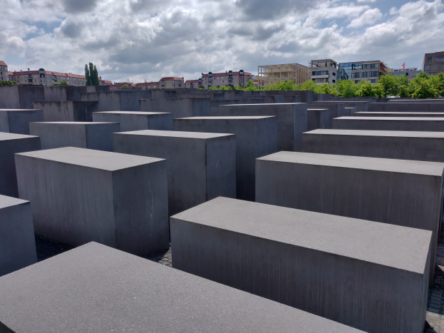 Konkreter Holocaust-Mahnmal zu den ermordeten Juden Europas mit rechteckigen Blöcken in einem Raster angeordnet, umgeben von Bäumen, Gebäuden und einem bewölkten Himmel.