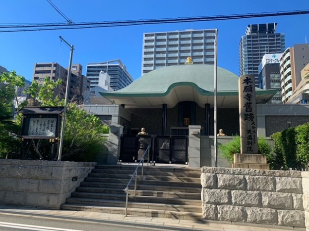 Großes Gebäude mit grüner Dachfläche in einer städtischen Umgebung mit umliegenden Treppen, Geländern, Beschilderung, Grünflächen und einer Straße darunter, vor einem Hintergrund aus Hochhäusern und einem klaren Himmel.