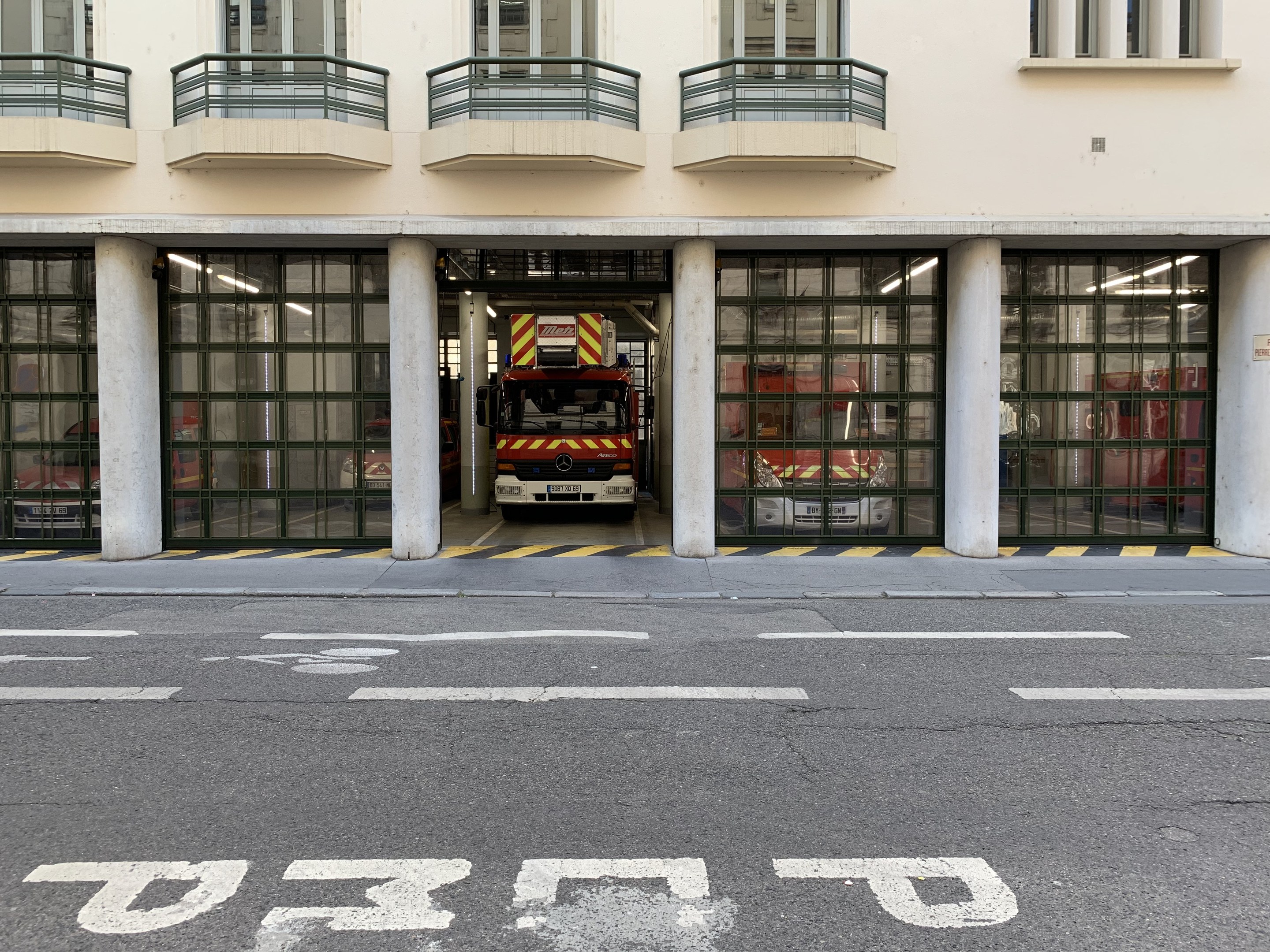 Feuerwache mit Feuerwehrauto davor, mit Fenstern, Geländern, Säulen und Glastüren, mit einer Straße unten.