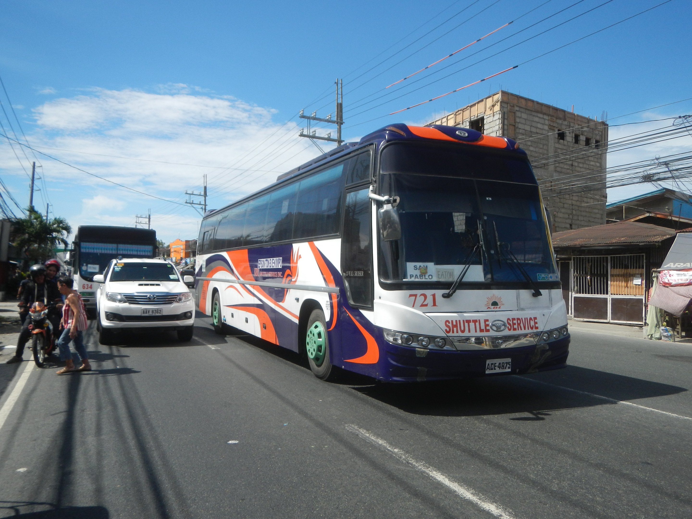 A shuttle bus drives down a street with tall buildings, electric poles with wires, and trees, while people walk on the footpath and a tent is set up nearby under a cloudy sky.