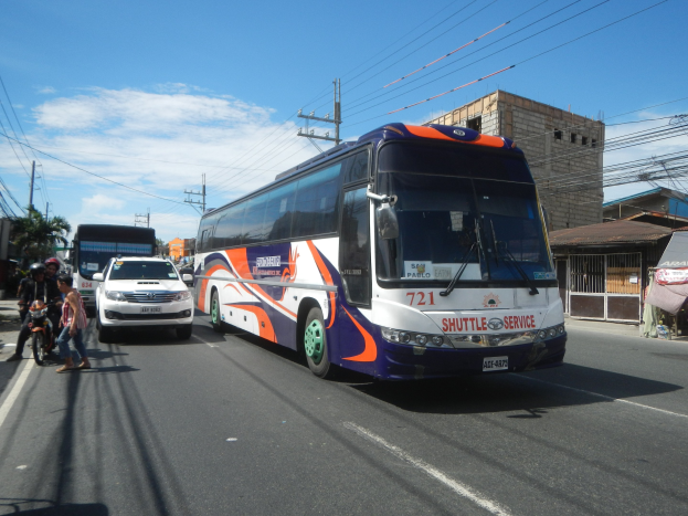 A shuttle bus drives down a street with tall buildings, electric poles with wires, and trees, while people walk on the footpath and a tent is set up nearby under a cloudy sky.