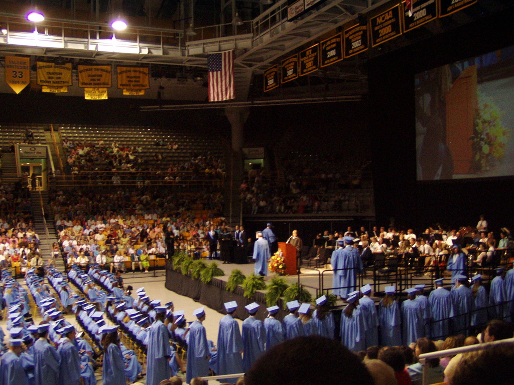 Eine Bühne mit Gruppen von Menschen, die sitzen und einige stehen, mit einem Podium mit Mikrofon und einem Blumenstrauß, Bannern, einer Flagge, einem Bildschirm, Treppen, leeren Stühlen und Scheinwerfern.