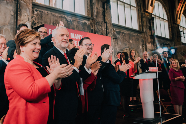 Eine Gruppe von Menschen, die vor einem Publikum stehen und jubeln, mit einem Podium, einem Mikrofon und einer Texttafel rechts sowie Stühlen, einer Fahne, einer Wand, Fenstern und Lichtern im Hintergrund.