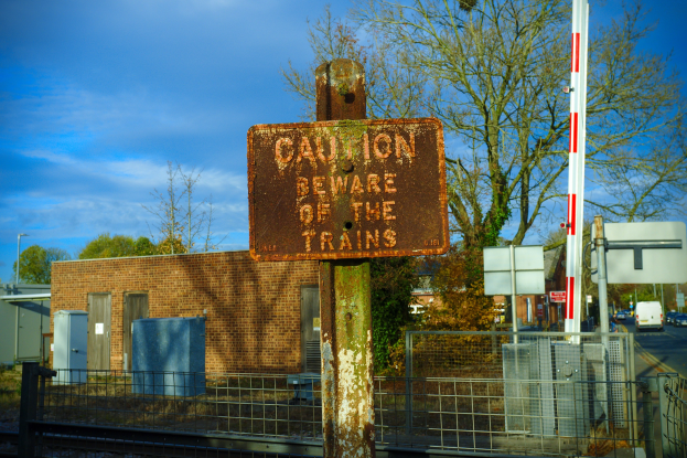 Vorsichtsschild auf einem Bahngeleise neben einem Metallzaun, mit Bäumen, Strommasten, einem Gebäude, Containern, Fahrzeugen auf einer Straße und einem bewölkten Himmel.