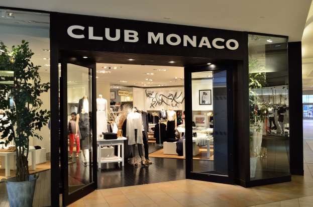 Außenansicht eines Club Monaco-Geschäfts in einem Einkaufszentrum mit Glaswänden und -türen, Schaufensterpuppen in Kleidern, Auslagetischen mit Artikeln, einem Topf mit einer Pflanze und Deckenbeleuchtung.