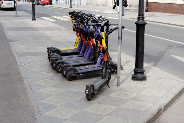 Eine Reihe von Elektro-Scootern, die an einer Stra√e mit fahrenden Fahrzeugen, Passanten auf dem Gehweg und Gebäuden im Hintergrund geparkt sind.