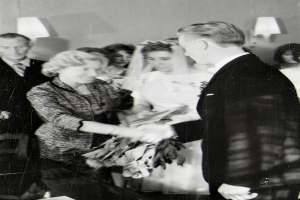 Ein Schwarz-Weiß-Foto eines Brautpaares beim Austausch der Eheringe, wobei die Braut in einem weißen Kleid und Schleier mit einem Strauß Blumen in der Hand zu sehen ist und der Bräutigam in einem Anzug und Krawatte steht, mit Wandlampen im Hintergrund.