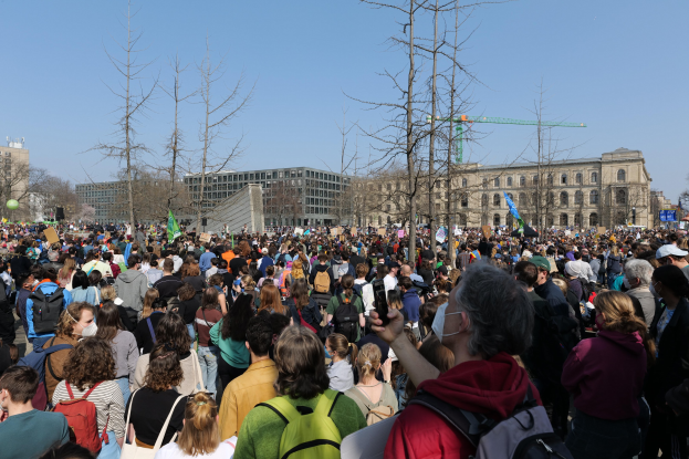 A large crowd of people stands in front of a building with windows, trees, and a clear sky, many holding placards and wearing bags, indicating a climate change protest.