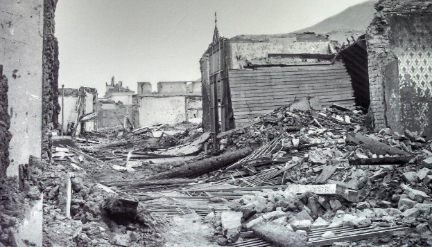 Schwarz-weiß-Foto eines schwer beschädigten Gebäudes auf einer Straße, mit Trümmern einschließlich eingestürzter Wände, Holzstöcken und Steinen, vor einem Hügel und einem Himmel