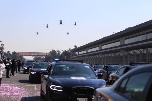 Polizeifahrzeuge vor einem Gebäude mit Menschen auf der linken Seite, Bäumen und einer Brücke im Hintergrund und vier Hubschraubern am Himmel.