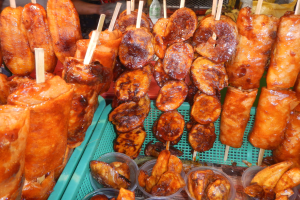 Marktstand mit Spießen von Hähnchenflügeln und verschiedenen Lebensmitteln in Plastikbehältern, mit Menschen und Flaschen im Hintergrund.