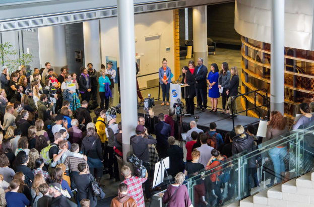 Eine Gruppe von Menschen steht auf einer Treppe, die zu einem Podium mit einem Mikrofon und Displaytafeln führt, und schaut auf eine Menge, die Kameras hält, bei einem Event mit Säulen, einer Tür und einem Topf mit einer Pflanze.