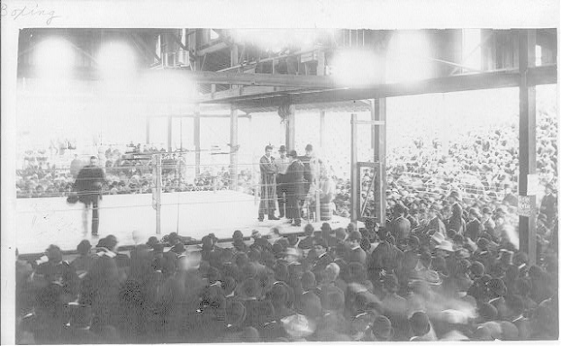 Schwarzes und weißes Foto einer Menge vor einer Bühne mit ein paar Menschen darauf und anderen im Publikum sitzend, was auf einen Boxkampf in einer Arena hinweist.