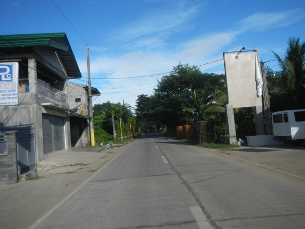 Leere Straße mit einem weißen Van auf der Seite geparkt, flankiert von verschlossenen Gebäuden mit einer Tafel mit Text, Bäumen, Strommasten, Drähten und einem klaren blauen Himmel im Hintergrund.