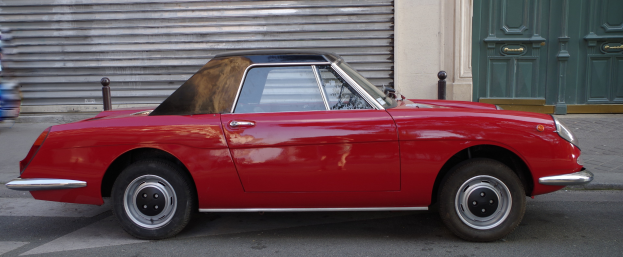 Rotes Auto auf der Straßenseite mit einer Person, die auf dem Gehweg links davon geht, Verschluss und grüne Tür im Hintergrund.