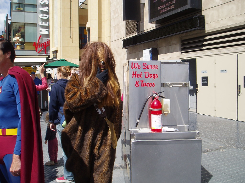 Eine Gruppe von Menschen, einige in Kostümen, versammelte sich vor Gebäuden mit sichtbarer Beschriftung, einer Box mit Schrift und einem Feuerlöscher in der Nähe.