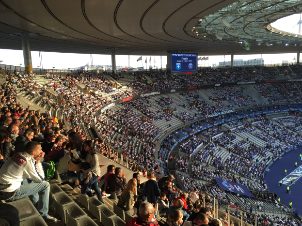 Große Menschenmenge in einem Stadion bei einem Füssballspiel mit einer Bühne, Fahnen, Masten und einem Bildschirm im Hintergrund
