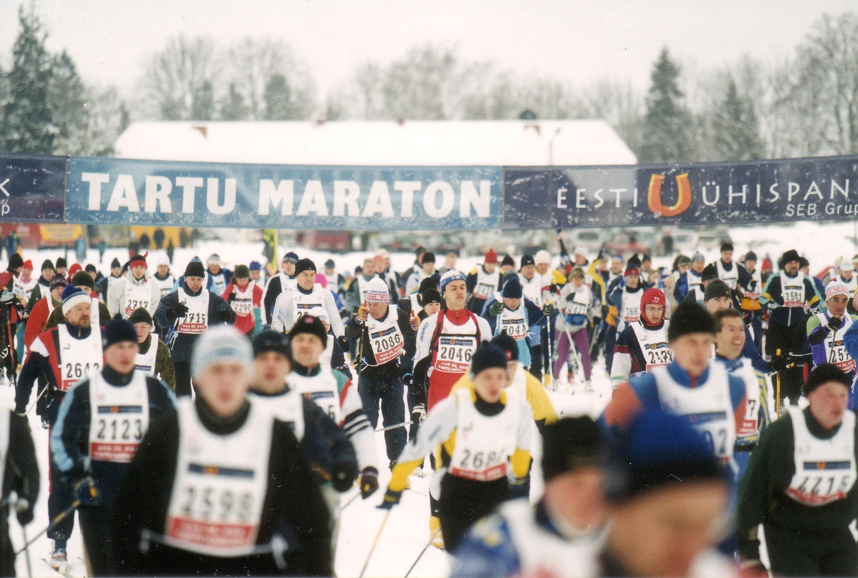 Group of people skiing in a race, wearing caps and holding ski poles, with banners, trees, and a clear blue sky in the background.