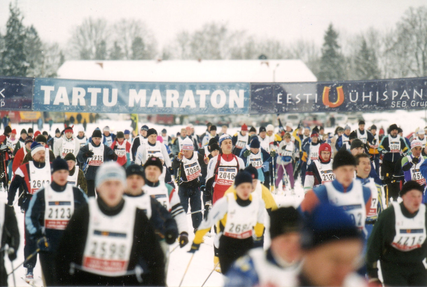 Group of people skiing in a race, wearing caps and holding ski poles, with banners, trees, and a clear blue sky in the background.
