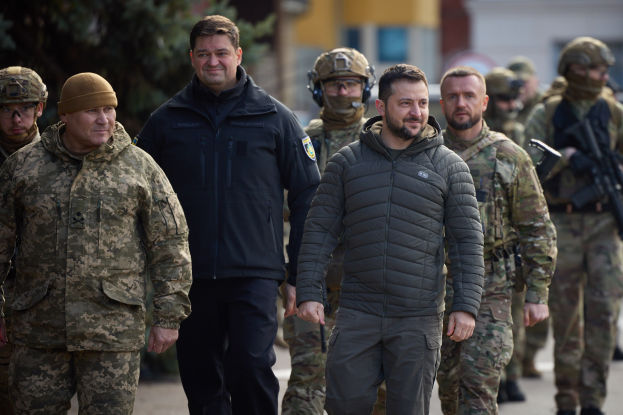 A group of men in military uniforms walking down a street, some wearing helmets and holding guns, with trees and buildings in the background.