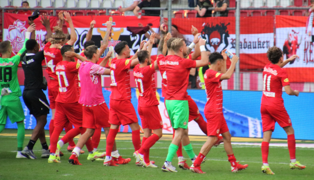 Group of soccer players celebrating a goal on a field, some holding bottles, others clapping, with banners, fencing, and stadium seating in the background.