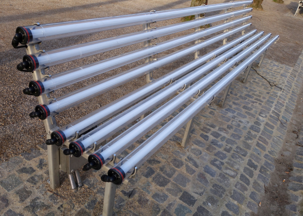 Eine Reihe von Solar-Wasserheizgeräten auf einem Kopfsteinpflasterweg unter einem klaren blauen Himmel, umgeben von Bäumen und hellem Sonnenlicht.