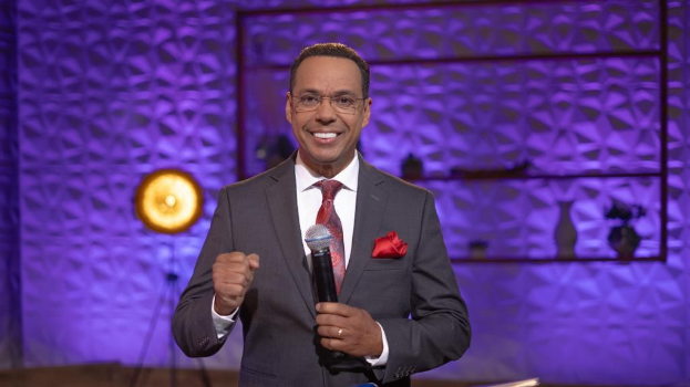 Stephen Colbert, Moderator von 'The Daily Show with Trevor Noah', steht auf einer Bühne und hält ein Mikrofon in der Hand, trägt einen Anzug, eine Krawatte und eine Brille, während im Hintergrund ein Licht und ein Ständer zu sehen sind.