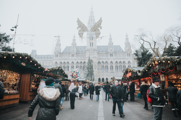 Ein lebendiger Weihnachtsmarkt in Wien, Österreich, mit Menschen, die herumlaufen, Ständen mit Lichtern und festlichen Décorations, einem Gebäude mit Fenstern im Hintergrund und einem klaren blauen Himmel.