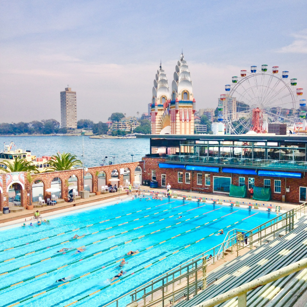 Large outdoor swimming pool with people swimming, surrounded by railings and benches, with a ferris wheel, buildings, trees, and cloudy sky in the background.