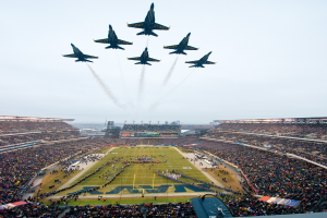 Eine Gruppe von Jets, die in Formation über ein Footballfeld fliegen, mit einem Stadion voller Menschen im Hintergrund.