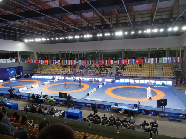 Großer Hallenstadium mit Zuschauern bei einem Wrestling-Kampf, einschließlich Sitzplätzen und Stehplätzen, Tischen mit Gegenständen und Hintergrundflaggen, Lichtern und Stangen.