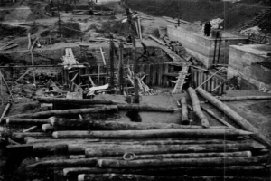 Ein Schwarz-Weiß-Foto einer Baustelle mit verstreuten Holzstämmen, Leitern, Pfählen und anderen Gegenständen, mit ein paar Menschen auf der rechten Seite und Bäumen im Hintergrund.