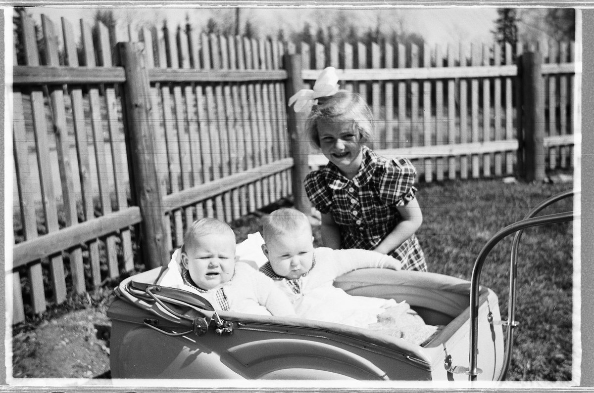 Schwarzes und weißes Foto von drei Kindern in einem Auto, mit einem Mädchen im Vordergrund, das steht und lächelt, einem Zaun und Bäumen im Hintergrund und grasbewachsenem Boden.
