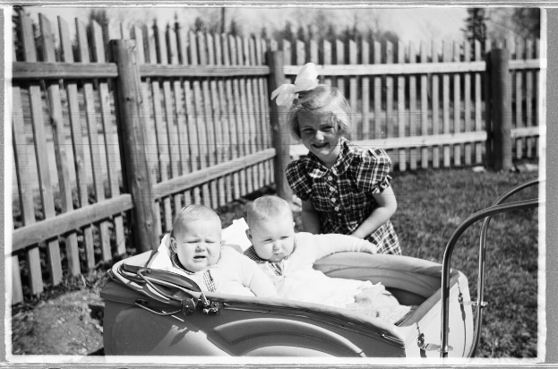 Schwarzes und weißes Foto von drei Kindern in einem Auto, mit einem Mädchen im Vordergrund, das steht und lächelt, einem Zaun und Bäumen im Hintergrund und grasbewachsenem Boden.