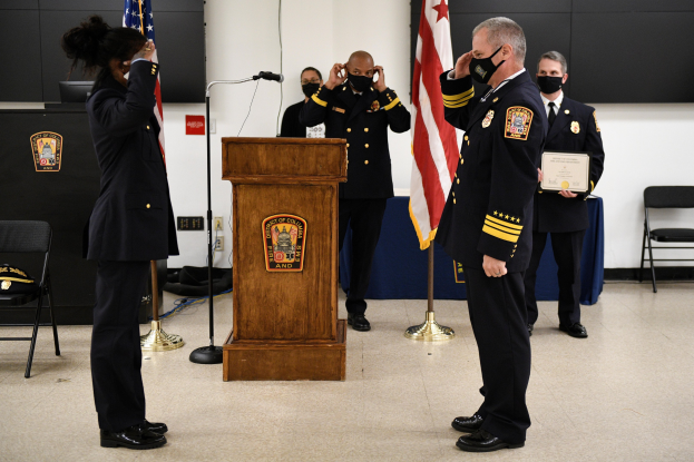Gruppe uniformierter Personen in Masken vor einem Podium mit Mikrofon, die eine Urkunde während einer Vereidigung eines neuen Feuerwehrchefs halten, umgeben von Fahnen, Stühlen, Tischen und einer Wand.