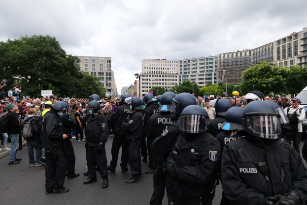 Eine große Gruppe von Polizisten steht vor einer Menschenmenge auf einer Straße, umgeben von Bäumen und Gebäuden unter einem bewölkten Himmel; einige in der Menge halten Kameras, was auf eine Demonstration in Berlin, Deutschland, hinweist.
