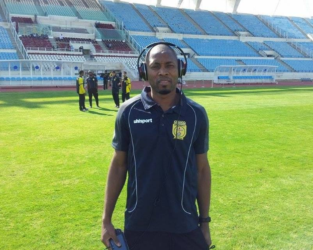 Ein Mann mit Headset, wahrscheinlich Trainer der ghanaischen Nationalmannschaft, steht auf einem Fussballfeld nähe eines Stadions, hält ein Objekt, während einige Personen im Hintergrund unter einem sichtbaren Himmel zu sehen sind.