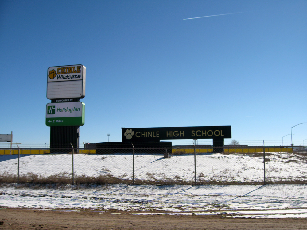 Eine Straße mit Schnee, ein Zaun mit Text, eine Wand, Laternenpfähle, Bäume und ein klarer Himmel, mit einem Schild im Vordergrund, auf dem "Chinle High School" steht.