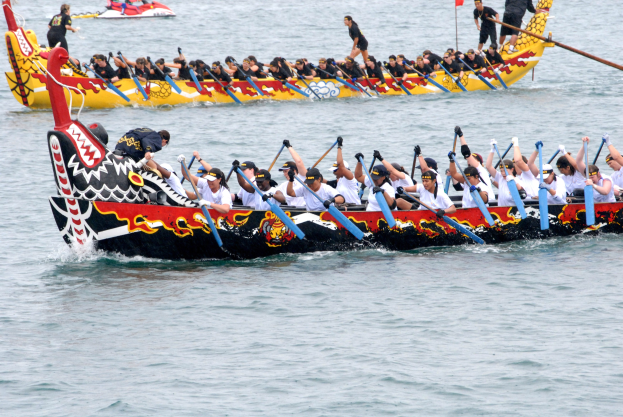 Group of people in a dragon boat racing on water, paddling, with another boat and a flag in the background.