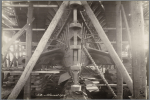 Ein Schwarz-Weiß-Foto eines Bootes in einem Schiffbau, umgeben von hölzernen Pfählen und anderen Gegenständen, mit Text unten.