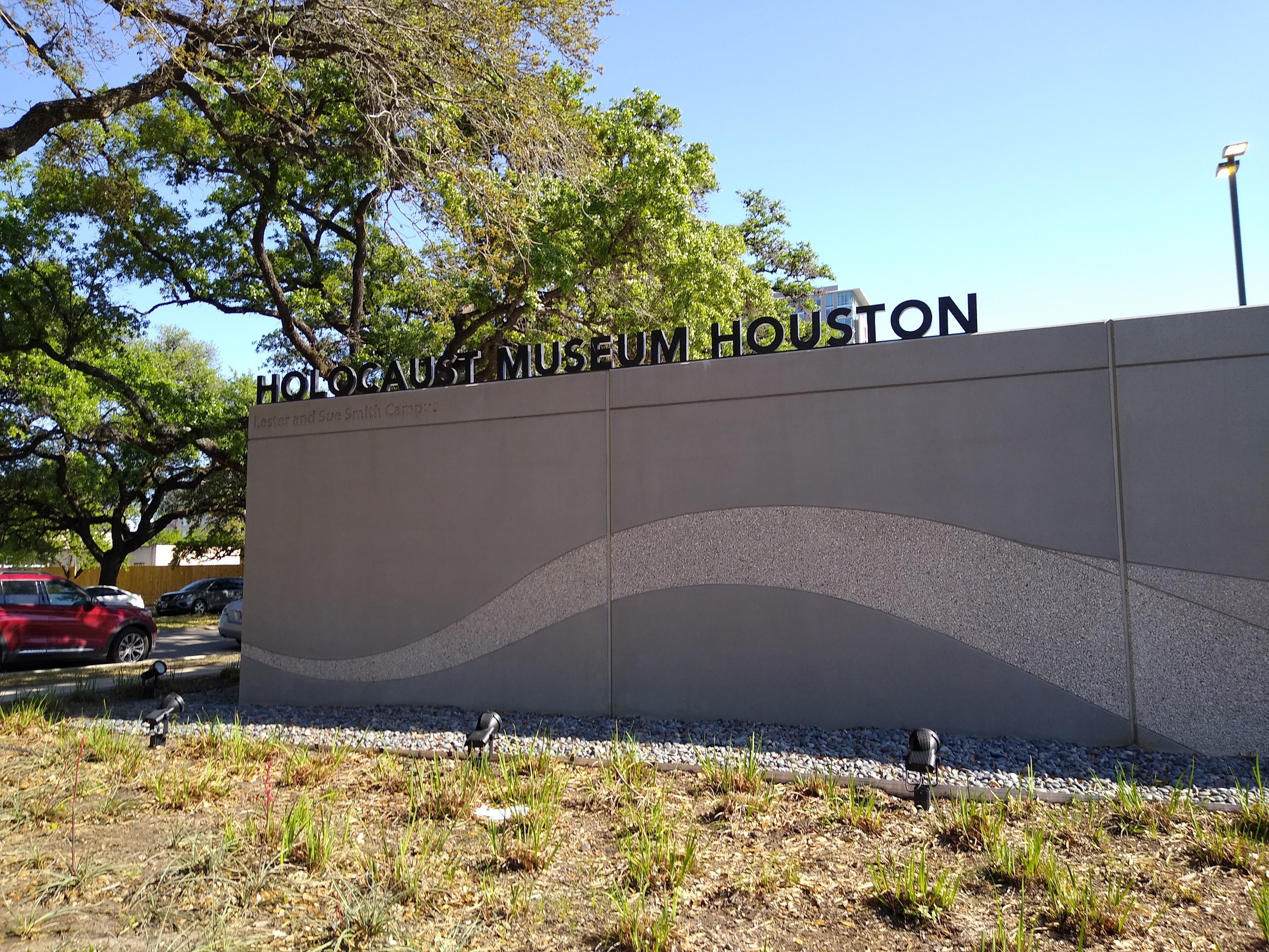 Außenansicht des Houstoner Holocaust-Museums mit einer strukturierten Wand, verstreuten Steinen, Grünfläche, Bäumen, vorbeifahrenden Fahrzeugen, einem Straßenpfahl und einem bewölkten Himmel.