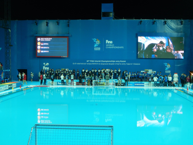 Eine Gruppe von Menschen steht um ein Hallenbad mit Netz am Boden herum, mit einer Wand im Hintergrund, auf der 'FINA World Championships 2015 Kazan' angezeigt wird, zusammen mit Trägern und Lampen.