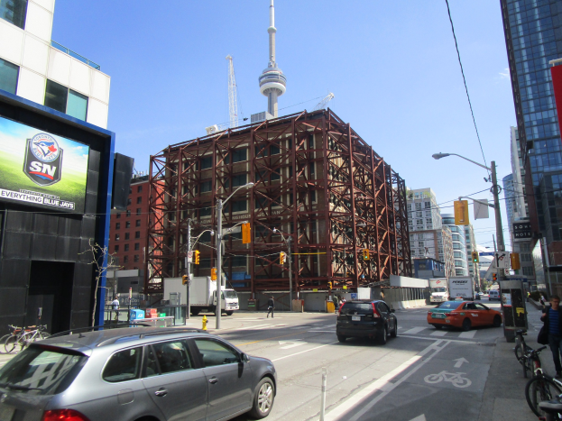Eine belebte Stadtstraße mit fahrenden Autos und Fahrrädern, Fußgängern auf dem Gehweg, einem großen Gebäude im Hintergrund, sichtbarer Straßeninfrastruktur und dem CN Tower in der Ferne unter einem klaren blauen Himmel.