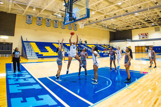 Eine Gruppe von Frauen, die Basketball in einer Turnhalle mit einem Scoreboard spielt, das ihren Sieg im NCAA-Turnier anzeigt, Zuschauern in den Rängen und Bannern an der Wand.