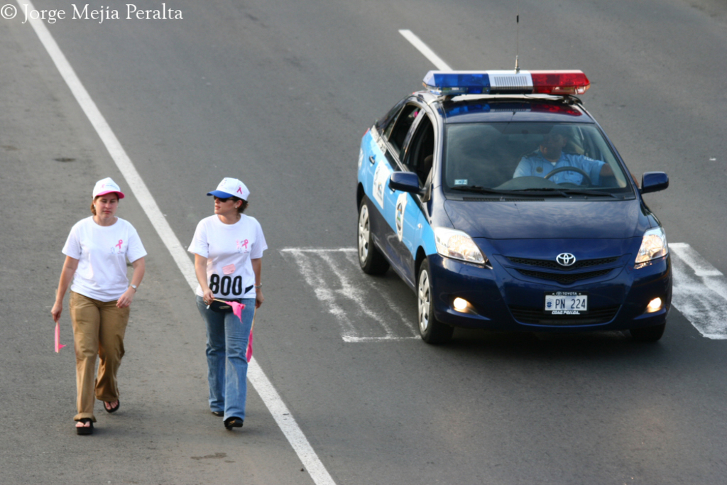 Zwei Frauen gehen auf einer Straße, eine hält ein rosa Papier und trägt eine rosa und weiße Mütze, die andere hält ein rosa Papier und trägt eine blaue und weiße Mütze, mit einem Streifenwagen im Hintergrund.