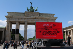A group of people stands before the Brandenburg Gate in Berlin, Germany, with a woman on a stage holding a microphone, a red sign reading "Speak Up to Commit to International Research Stations in Antarctica," and trees and clouds in the background.